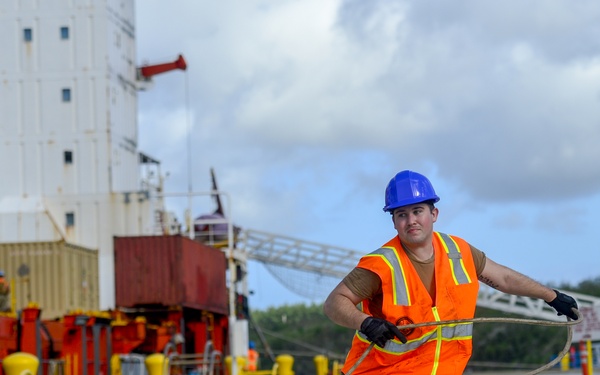 A worker in military-style camouflage pants and a high-visibility orange safety vest is pulling a thick mooring rope on a port or naval pier. He is wearing a blue hard hat and black gloves, indicating active dock work near a large vessel. The setting appears to be a military or commercial port facility with shipping containers and dock equipment visible in the background.
