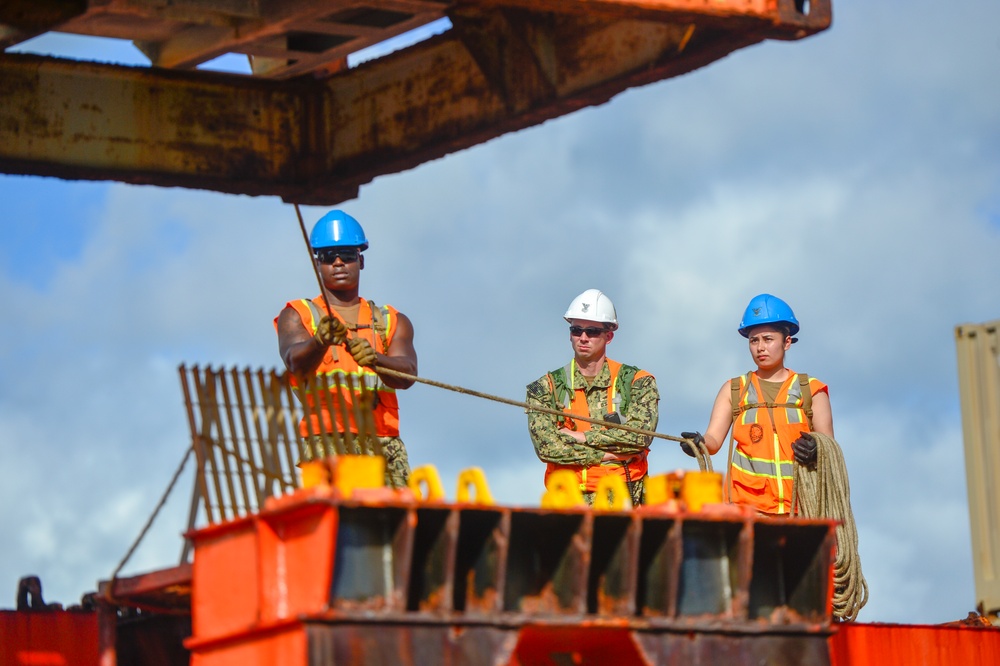 Three workers are positioned on what appears to be a large orange vessel or maritime structure, engaged in rope/line handling operations. Two workers wear blue hard hats and orange high-visibility safety vests, while the center individual wears a white hard hat and military-style camouflage uniform with an orange safety vest. The setting appears to be a port or harbor environment with partially cloudy skies visible in the background.