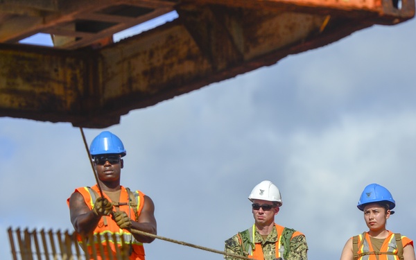 Three workers are positioned on what appears to be a large orange vessel or maritime structure, engaged in rope/line handling operations. Two workers wear blue hard hats and orange high-visibility safety vests, while the center individual wears a white hard hat and military-style camouflage uniform with an orange safety vest. The setting appears to be a port or harbor environment with partially cloudy skies visible in the background.