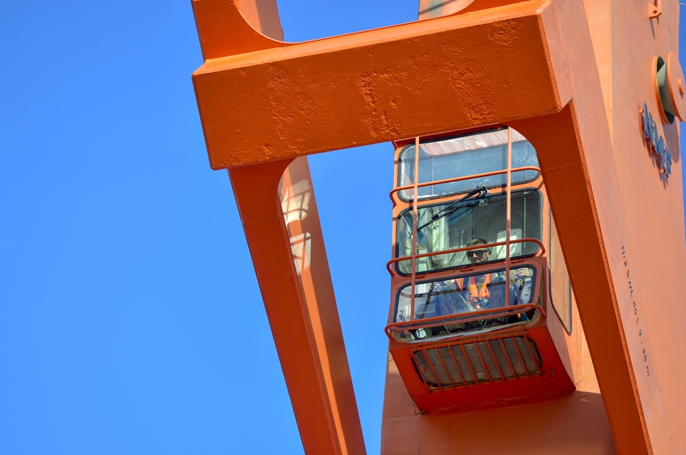 The image shows a close-up, low-angle view of an operator's cab mounted on a large orange industrial crane or port gantry structure. A worker wearing high-visibility orange safety gear is visible inside the enclosed glass cab. The photo is taken against a clear, cloudless blue sky.