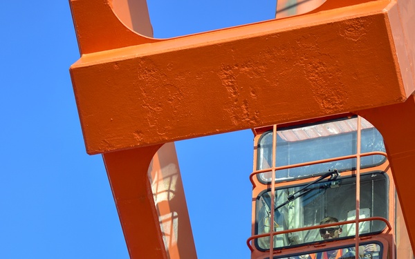 The image shows a close-up, low-angle view of an operator's cab mounted on a large orange industrial crane or port gantry structure. A worker wearing high-visibility orange safety gear is visible inside the enclosed glass cab. The photo is taken against a clear, cloudless blue sky.