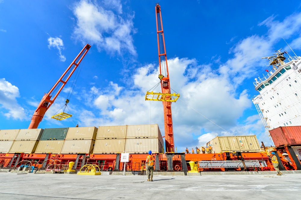 A busy port/dock scene showing cargo operations with two large orange cranes actively lifting shipping containers from a vessel. Multiple workers in high-visibility gear are visible on the dock and ship deck, overseeing the unloading process. The scene is photographed from a low angle against a bright blue sky with scattered clouds.