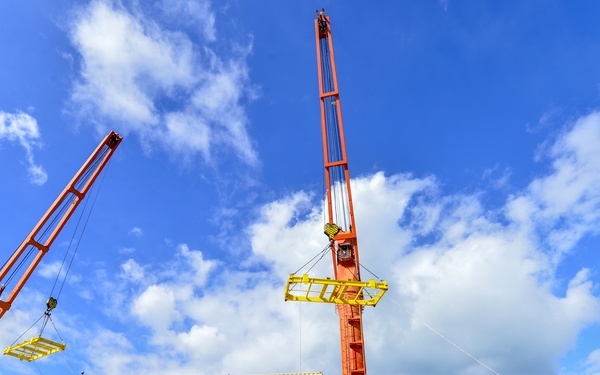 A busy port/dock scene showing cargo operations with two large orange cranes actively lifting shipping containers from a vessel. Multiple workers in high-visibility gear are visible on the dock and ship deck, overseeing the unloading process. The scene is photographed from a low angle against a bright blue sky with scattered clouds.
