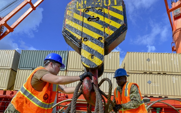Two workers in military-style camouflage uniforms, orange high-visibility safety vests, and blue hard hats are operating rigging equipment at a port or shipyard. They are attaching or managing heavy wire rope cables connected to a large crane hook marked with black and yellow hazard stripes. The background shows stacked shipping containers and red port cranes under a partly cloudy blue sky.