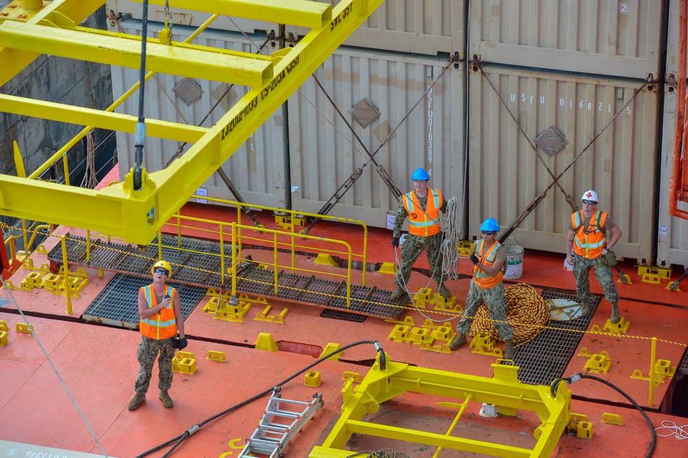 Four military personnel in Navy camouflage uniforms and high-visibility orange safety vests are working on the deck of a cargo or military vessel. They are engaged in deck operations involving ropes and rigging near yellow safety structures and shipping containers. One individual wears a white hard hat with a red cross symbol, suggesting a medical or safety officer role.