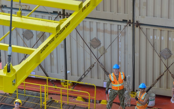 Four military personnel in Navy camouflage uniforms and high-visibility orange safety vests are working on the deck of a cargo or military vessel. They are engaged in deck operations involving ropes and rigging near yellow safety structures and shipping containers. One individual wears a white hard hat with a red cross symbol, suggesting a medical or safety officer role.