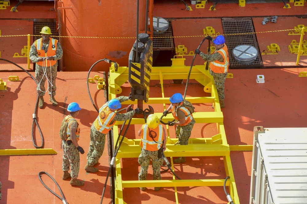 A group of military personnel, likely U.S. Navy sailors, are working on the deck of a large orange vessel or ship. They are handling heavy wire rope/cable rigging connected to a yellow lifting frame or spreader bar. One individual in a yellow hard hat appears to be in a supervisory role while the others actively manage the rigging equipment.