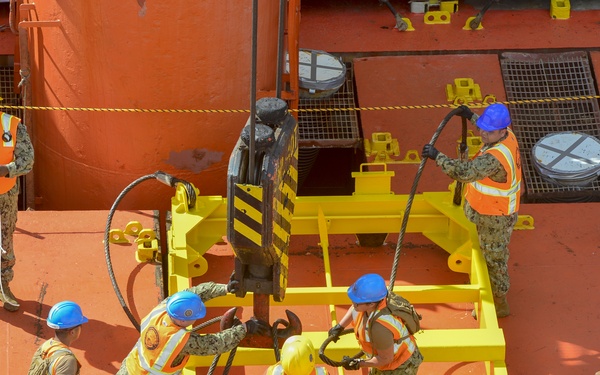 A group of military personnel, likely U.S. Navy sailors, are working on the deck of a large orange vessel or ship. They are handling heavy wire rope/cable rigging connected to a yellow lifting frame or spreader bar. One individual in a yellow hard hat appears to be in a supervisory role while the others actively manage the rigging equipment.