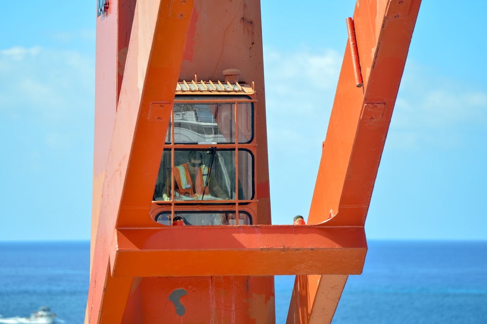 A worker wearing an orange high-visibility vest is visible inside the operator cab of a large orange crane or heavy lifting structure. The equipment appears to be located near or on a marine vessel, with open ocean and blue sky visible in the background. A second person's head is partially visible at the lower right of the frame.