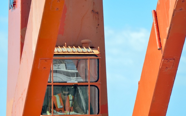 A worker wearing an orange high-visibility vest is visible inside the operator cab of a large orange crane or heavy lifting structure. The equipment appears to be located near or on a marine vessel, with open ocean and blue sky visible in the background. A second person's head is partially visible at the lower right of the frame.