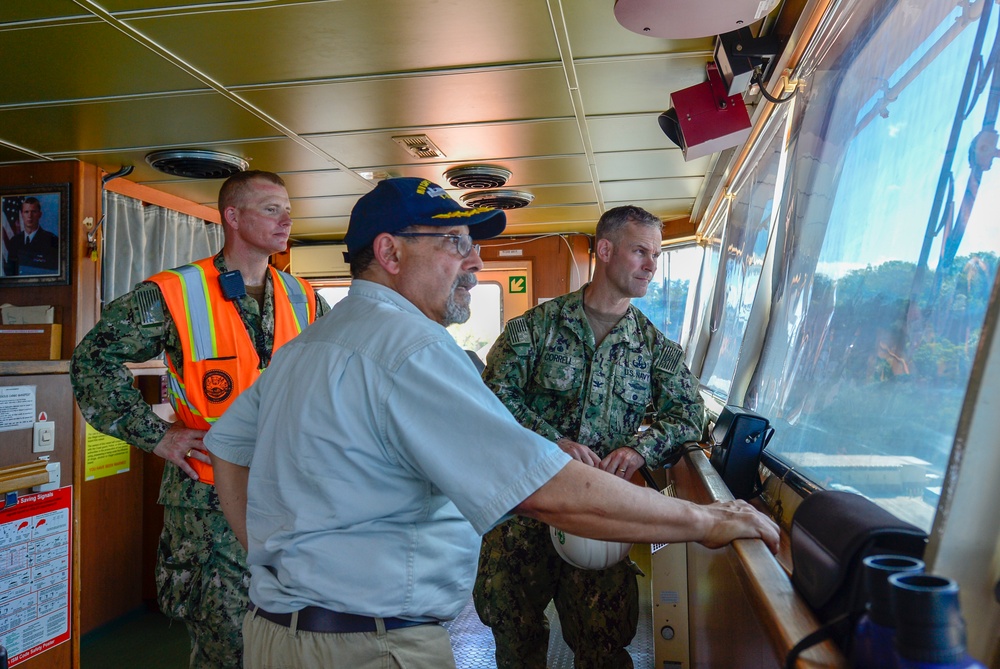 Three individuals are gathered on the bridge/wheelhouse of a naval or military vessel. Two are wearing U.S. Navy camouflage uniforms, one of whom also has an orange high-visibility safety vest, while a third civilian-dressed man wearing a navy blue cap leans toward the forward windows. They appear to be reviewing operations or conducting an inspection of the vessel's bridge.