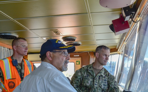 Three individuals are gathered on the bridge/wheelhouse of a naval or military vessel. Two are wearing U.S. Navy camouflage uniforms, one of whom also has an orange high-visibility safety vest, while a third civilian-dressed man wearing a navy blue cap leans toward the forward windows. They appear to be reviewing operations or conducting an inspection of the vessel's bridge.