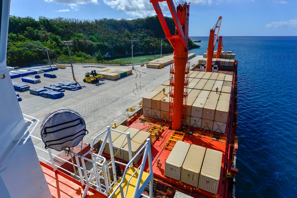 An aerial view from the bridge or upper deck of a large container ship docked at a port, showing the vessel's deck loaded with stacked shipping containers in beige and red colors. The port facility is visible alongside, featuring a wide concrete apron, a yellow forklift in operation, and stacked containers on the quayside. A lush green hillside and calm blue ocean form the backdrop, suggesting a tropical island port location.