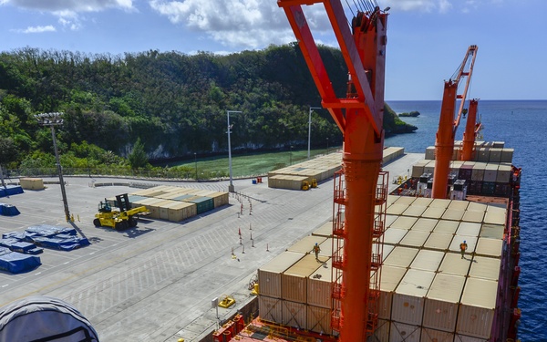 An aerial view from the bridge or upper deck of a large container ship docked at a port, showing the vessel's deck loaded with stacked shipping containers in beige and red colors. The port facility is visible alongside, featuring a wide concrete apron, a yellow forklift in operation, and stacked containers on the quayside. A lush green hillside and calm blue ocean form the backdrop, suggesting a tropical island port location.