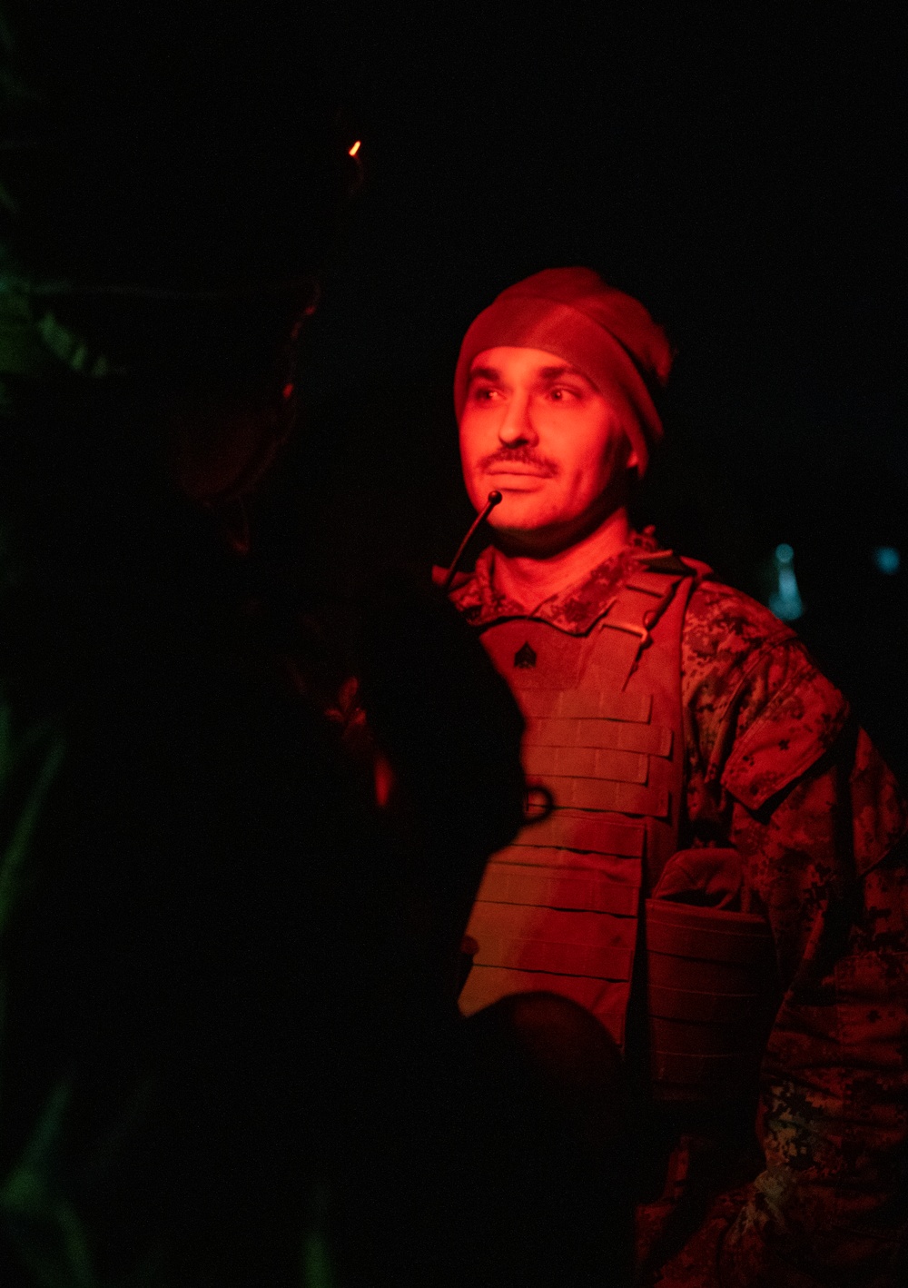 A male soldier is photographed at night, illuminated by red light, wearing tactical gear including a plate carrier vest and camouflage uniform. He has a communication headset/microphone near his mouth and is wearing a beanie-style cap. A small light source is visible in the upper background.