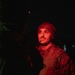 A male soldier is photographed at night, illuminated by red light, wearing tactical gear including a plate carrier vest and camouflage uniform. He has a communication headset/microphone near his mouth and is wearing a beanie-style cap. A small light source is visible in the upper background.