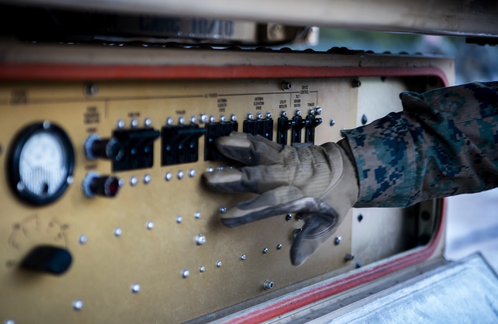 A person wearing military digital camouflage (MARPAT pattern) uniform and tan tactical gloves is operating a control panel on what appears to be military equipment. The panel contains multiple circuit breakers, toggle switches, indicator lights, and labeled sections including 'AC Power,' 'Antenna Elevation Drive,' 'Utility Outlets,' 'Sensor Heater,' and 'Trailer' controls. The image is taken in close-up with shallow depth of field, emphasizing the hand interaction with the panel.