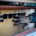 A person wearing military digital camouflage (MARPAT pattern) uniform and tan tactical gloves is operating a control panel on what appears to be military equipment. The panel contains multiple circuit breakers, toggle switches, indicator lights, and labeled sections including 'AC Power,' 'Antenna Elevation Drive,' 'Utility Outlets,' 'Sensor Heater,' and 'Trailer' controls. The image is taken in close-up with shallow depth of field, emphasizing the hand interaction with the panel.