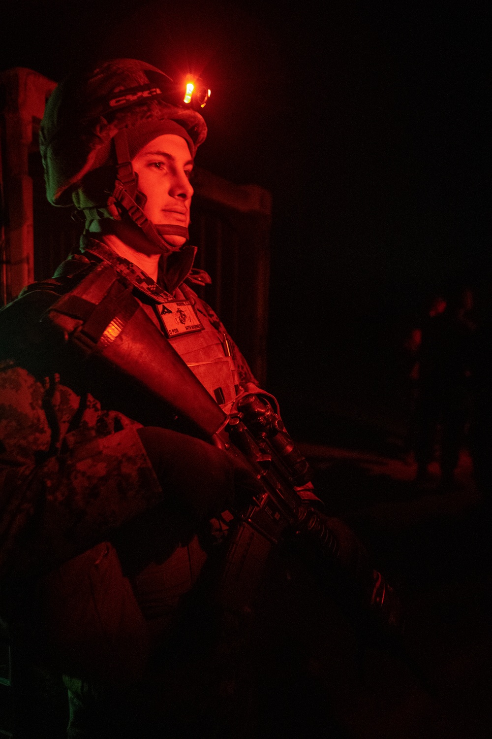 A military service member is photographed at night under red light illumination, wearing full combat gear including a tactical helmet with a red indicator light. The individual appears to be a U.S. Marine based on the visible Eagle, Globe, and Anchor emblem patch on their body armor. The red lighting creates a dramatic, low-light operational atmosphere consistent with night operations.