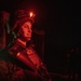 A military service member is photographed at night under red light illumination, wearing full combat gear including a tactical helmet with a red indicator light. The individual appears to be a U.S. Marine based on the visible Eagle, Globe, and Anchor emblem patch on their body armor. The red lighting creates a dramatic, low-light operational atmosphere consistent with night operations.