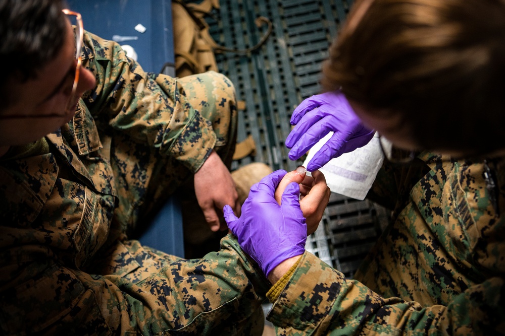 Two military personnel in U.S. Marine Corps MARPAT digital camouflage uniforms are engaged in a medical treatment procedure. One individual wearing purple nitrile gloves and a face mask is treating what appears to be a hand or foot injury on the other service member. The setting appears to be inside a military vehicle or aircraft based on the metal grating visible in the background.