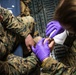 Two military personnel in U.S. Marine Corps MARPAT digital camouflage uniforms are engaged in a medical treatment procedure. One individual wearing purple nitrile gloves and a face mask is treating what appears to be a hand or foot injury on the other service member. The setting appears to be inside a military vehicle or aircraft based on the metal grating visible in the background.