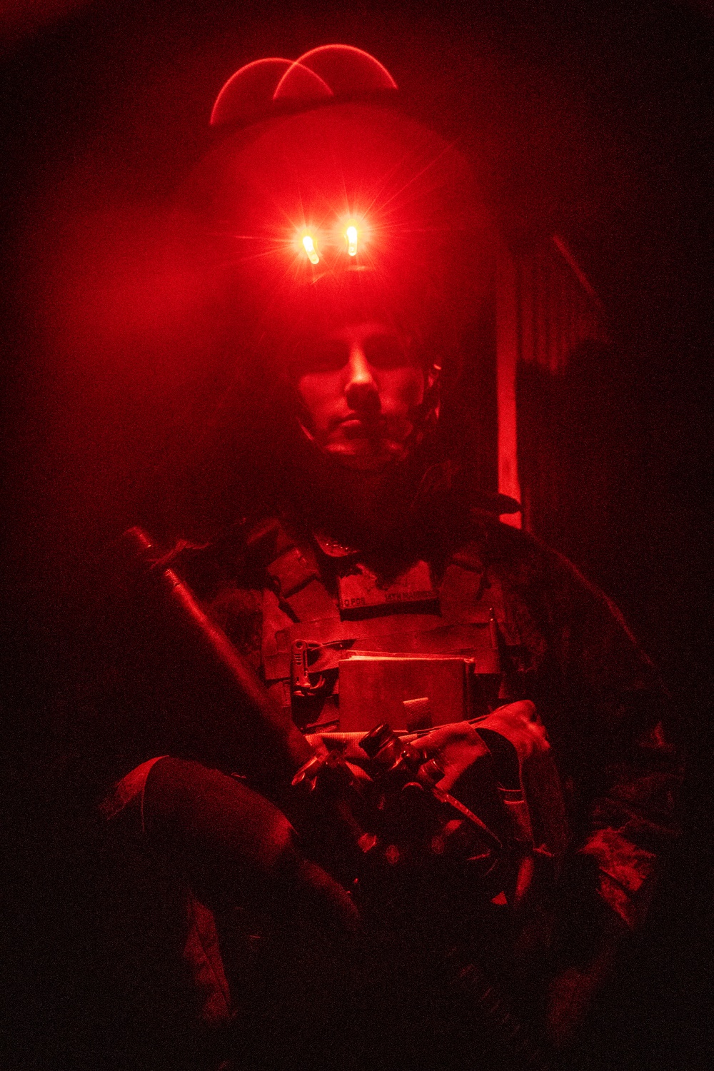 A military or tactical operator is photographed under intense red lighting in a dark environment. The subject is wearing full tactical gear including body armor and is holding a weapon, with what appears to be night-vision device mounts on their helmet emitting red light. The dramatic red illumination creates a high-contrast, atmospheric image with significant shadow detail loss.