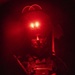 A military or tactical operator is photographed under intense red lighting in a dark environment. The subject is wearing full tactical gear including body armor and is holding a weapon, with what appears to be night-vision device mounts on their helmet emitting red light. The dramatic red illumination creates a high-contrast, atmospheric image with significant shadow detail loss.