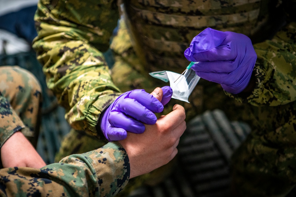 A military medic wearing purple nitrile gloves is preparing to administer an injection or perform a medical procedure on another service member's hand or wrist. Both individuals are wearing camouflage uniforms consistent with U.S. Marine Corps MARPAT pattern. The medic is removing a needle/syringe from its sterile packaging.