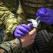A military medic wearing purple nitrile gloves is preparing to administer an injection or perform a medical procedure on another service member's hand or wrist. Both individuals are wearing camouflage uniforms consistent with U.S. Marine Corps MARPAT pattern. The medic is removing a needle/syringe from its sterile packaging.