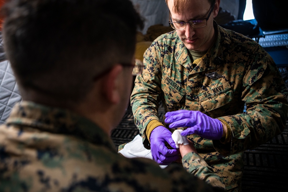 A U.S. Navy corpsman wearing MARPAT camouflage uniform is performing a medical procedure on another service member's arm or hand. The corpsman, whose name tape reads 'ROSSELL' and whose uniform is labeled 'U.S. NAVY', is wearing purple nitrile gloves and appears to be drawing blood or administering an injection. The setting appears to be a field medical environment or temporary medical station.