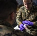 A U.S. Navy corpsman wearing MARPAT camouflage uniform is performing a medical procedure on another service member's arm or hand. The corpsman, whose name tape reads 'ROSSELL' and whose uniform is labeled 'U.S. NAVY', is wearing purple nitrile gloves and appears to be drawing blood or administering an injection. The setting appears to be a field medical environment or temporary medical station.
