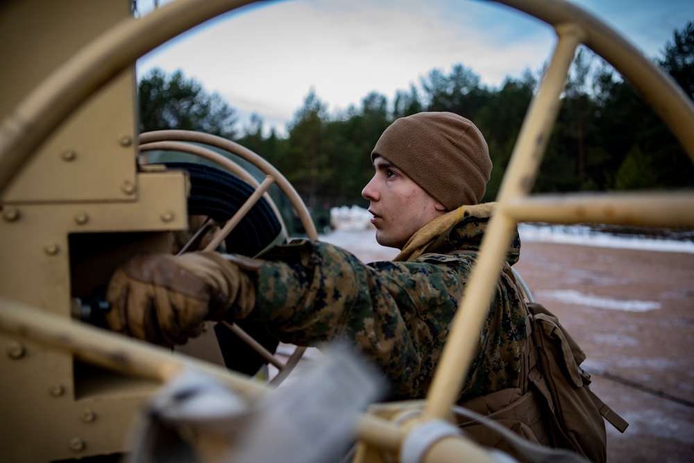 A young military service member in MARPAT camouflage uniform and brown fleece cap is operating or adjusting equipment on what appears to be a military vehicle. The photo is taken through a large steering wheel or mechanical frame, creating a framing effect. The setting appears to be an outdoor training environment with snow-dusted ground and pine trees in the background.