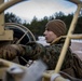 A young military service member in MARPAT camouflage uniform and brown fleece cap is operating or adjusting equipment on what appears to be a military vehicle. The photo is taken through a large steering wheel or mechanical frame, creating a framing effect. The setting appears to be an outdoor training environment with snow-dusted ground and pine trees in the background.
