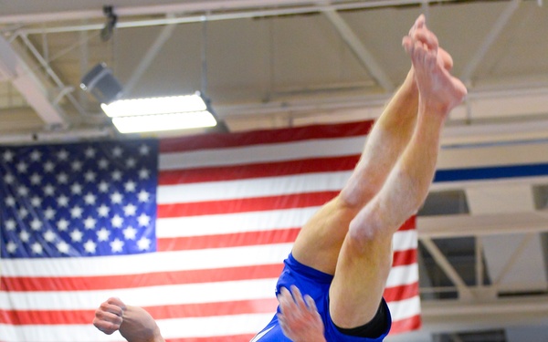 A male gymnast wearing a blue 'Air Force' uniform is captured mid-air performing an inverted flip or back layout in an indoor gymnasium. An American flag is prominently displayed in the background, and spectators in blue seating are visible below. The image is taken from a low angle, emphasizing the athlete's height and body position during the skill.