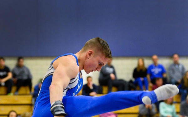 A male gymnast wearing a blue competition leotard performs a routine on the pommel horse during an indoor gymnastics meet. He is captured mid-movement with one leg extended high in the air while gripping the pommels. Spectators are seated in wooden bleachers in the background of what appears to be a gymnasium.