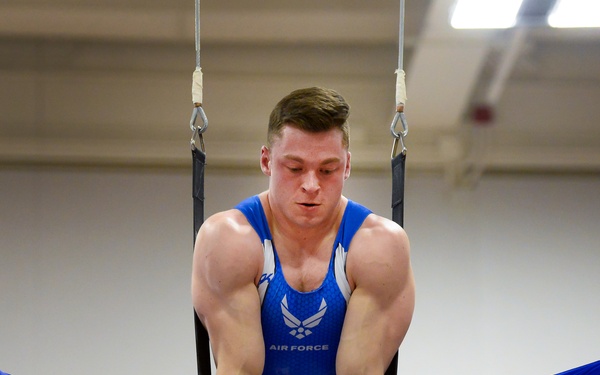 A male gymnast wearing a blue 'Air Force' uniform is performing on still rings in an indoor gymnasium. He is executing a straddle hold position with his legs spread wide apart while gripping the rings with both hands. The athlete displays significant upper body musculature and is wearing wrist wraps with grip supports.