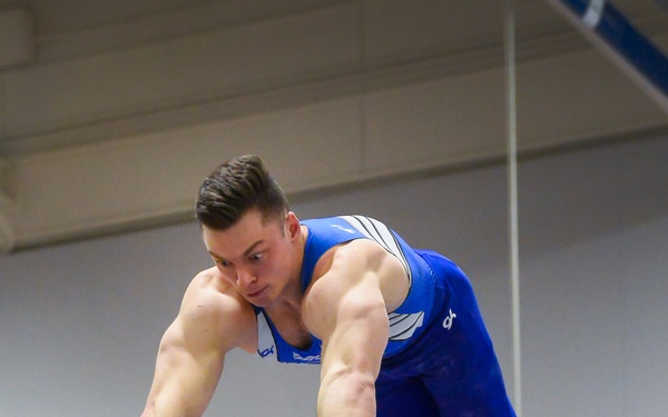 A male gymnast is performing on the horizontal bar (high bar) in an indoor gymnasium. He is captured mid-routine, body nearly parallel to the bar with arms extended and gripping the apparatus. He wears a blue competition leotard, gymnastics grips with green wrist straps, and white socks.