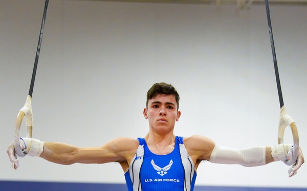 A male gymnast wearing a U.S. Air Force uniform performs on the still rings, executing an iron cross or similar static hold with arms fully extended horizontally. He is suspended in mid-air in a gymnasium displaying AFA (Air Force Academy) Men's Gymnastics championship banners on the wall behind him. The athlete wears gymnastics grips/hand guards and full blue competition attire.