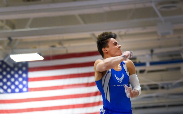 A male gymnast wearing a U.S. Air Force uniform is captured mid-air above a pommel horse or vault apparatus during a gymnastics competition. He is performing in what appears to be an indoor gymnasium decorated with an American flag and blue team colors. Spectators and teammates in blue uniforms are visible in the background.