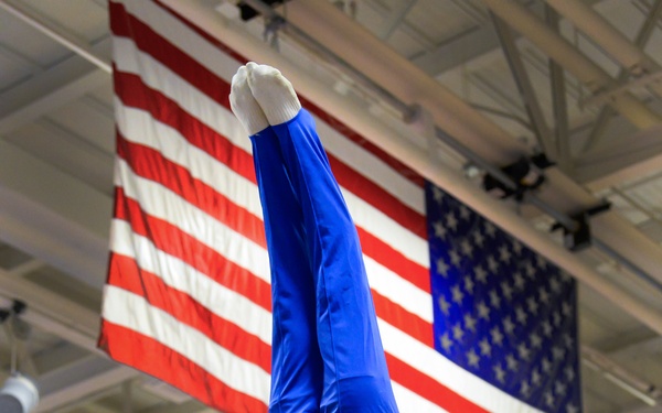 A male gymnast wearing a blue 'Air Force' uniform performs a handstand on parallel bars in an indoor gymnasium. He is inverted with legs pointing straight up, demonstrating exceptional strength and balance. Two large American flags are prominently displayed hanging from the ceiling in the background.