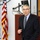 A middle-aged man in a dark business suit and striped tie is speaking or presenting at a podium in what appears to be an official or government venue. He is standing near a wooden lectern with a monitor mounted above it, with an American flag visible to his left. The setting suggests a formal briefing, lecture, or official presentation.