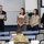 A formal presentation or award ceremony is taking place in what appears to be a military or government classroom setting. An older man in a suit is reading from a framed certificate or plaque while three women stand beside him. The audience includes at least one person in military camouflage uniform.