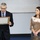 Two individuals stand in front of a whiteboard in what appears to be a classroom or meeting room setting. An older man in a dark suit is reading from a framed plaque or certificate he is holding, while a younger woman in a gold/bronze velvet top stands beside him with her hands clasped. The scene suggests a formal recognition or award presentation ceremony.