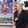 A middle-aged man in a dark suit and striped tie is speaking or presenting at what appears to be a military or government briefing. He stands in front of a display banner featuring National Guard insignia and imagery, alongside an American flag. The setting suggests an official military or governmental presentation environment.