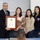 Four individuals are posing together in what appears to be a classroom or meeting room setting during a formal award ceremony. They are holding a framed citation from the State of Texas, suggesting an official recognition or honor is being presented. The group consists of one older man in a suit and three younger women, one of whom is wearing a letterman-style jacket with Texas-related patches.