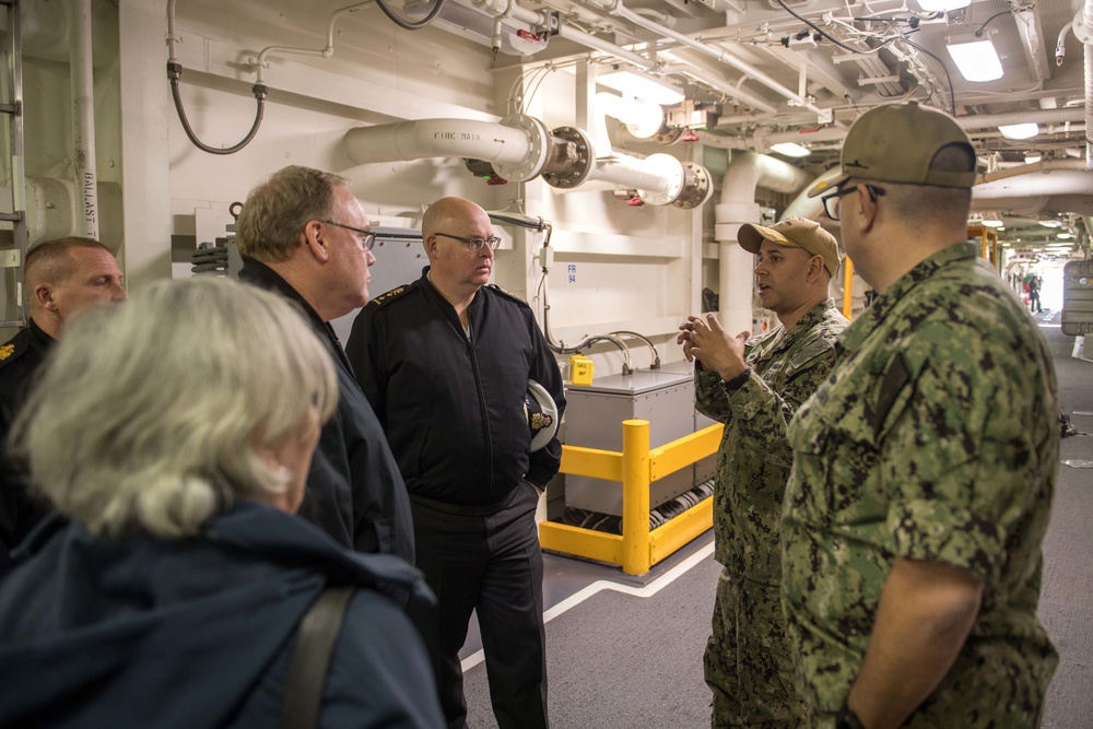 A group of military and civilian personnel are gathered inside what appears to be the interior deck of a naval vessel. Two U.S. Navy sailors in digital camouflage (NWU Type III) uniforms are briefing or explaining something to a group that includes officers in dark naval uniforms and at least one civilian. The setting shows the utilitarian interior of a warship with visible piping, conduits, and safety markings.