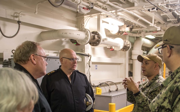 A group of military and civilian personnel are gathered inside what appears to be the interior deck of a naval vessel. Two U.S. Navy sailors in digital camouflage (NWU Type III) uniforms are briefing or explaining something to a group that includes officers in dark naval uniforms and at least one civilian. The setting shows the utilitarian interior of a warship with visible piping, conduits, and safety markings.