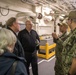 A group of military and civilian personnel are gathered inside what appears to be the interior deck of a naval vessel. Two U.S. Navy sailors in digital camouflage (NWU Type III) uniforms are briefing or explaining something to a group that includes officers in dark naval uniforms and at least one civilian. The setting shows the utilitarian interior of a warship with visible piping, conduits, and safety markings.