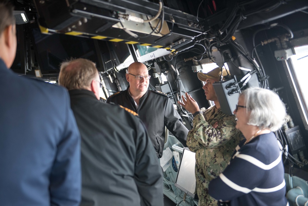 A group of individuals, including at least one person in US Navy camouflage uniform and others in dark naval or civilian attire, are gathered inside what appears to be a military vessel's bridge or command area. A uniformed service member appears to be briefing or explaining something to the group, gesturing with their hand. The setting is characterized by overhead equipment, cables, and yellow hazard markings typical of a warship interior.